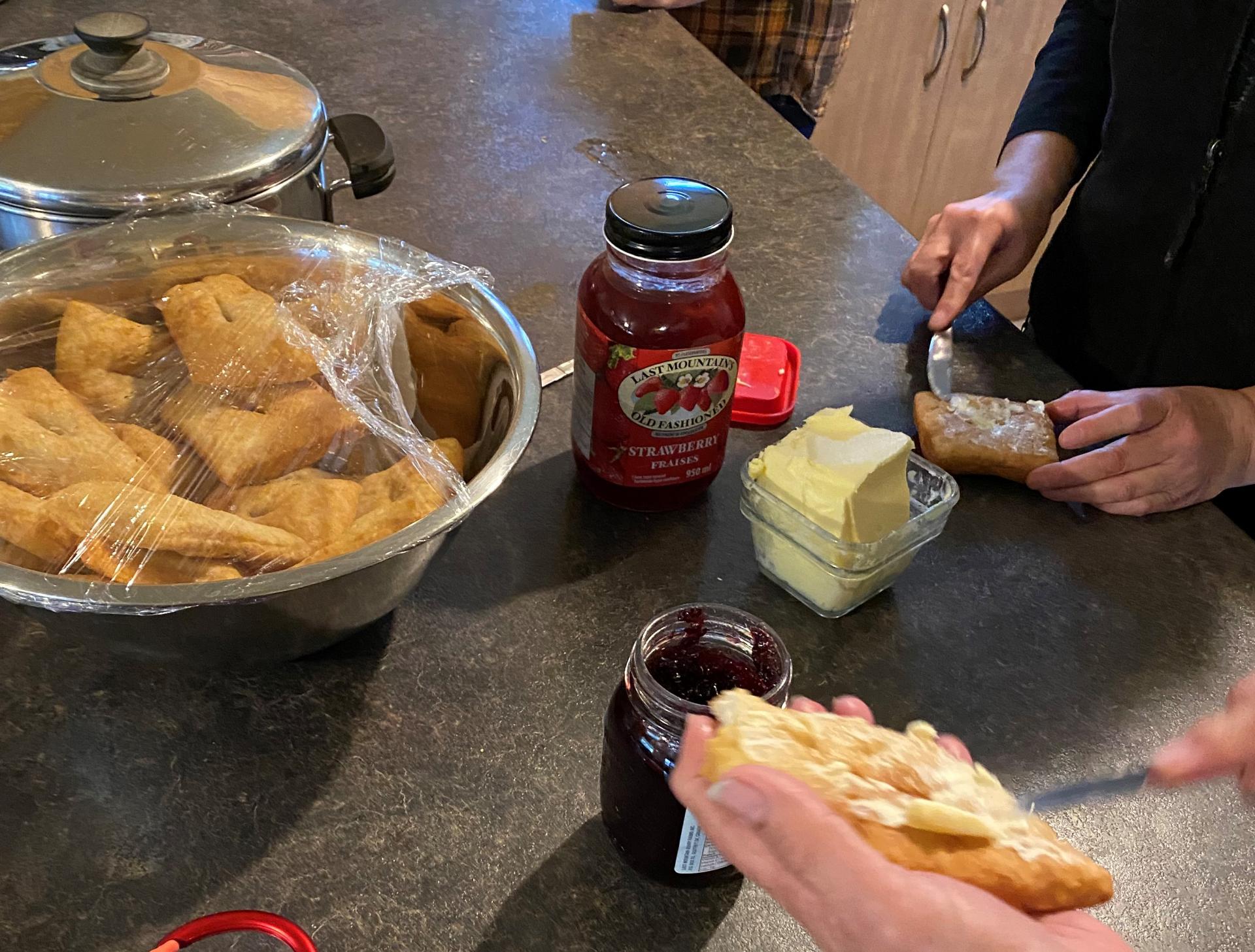 Close up of two women's hands spreading butter on fried bannock, with a bowl of bannock and jam on the counter