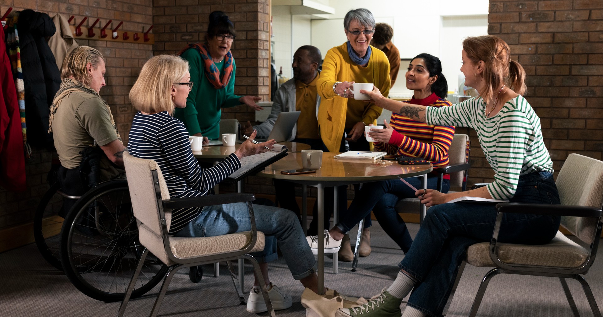 Diverse group of people meet in a community centre around a table. They are smiling and getting work done.