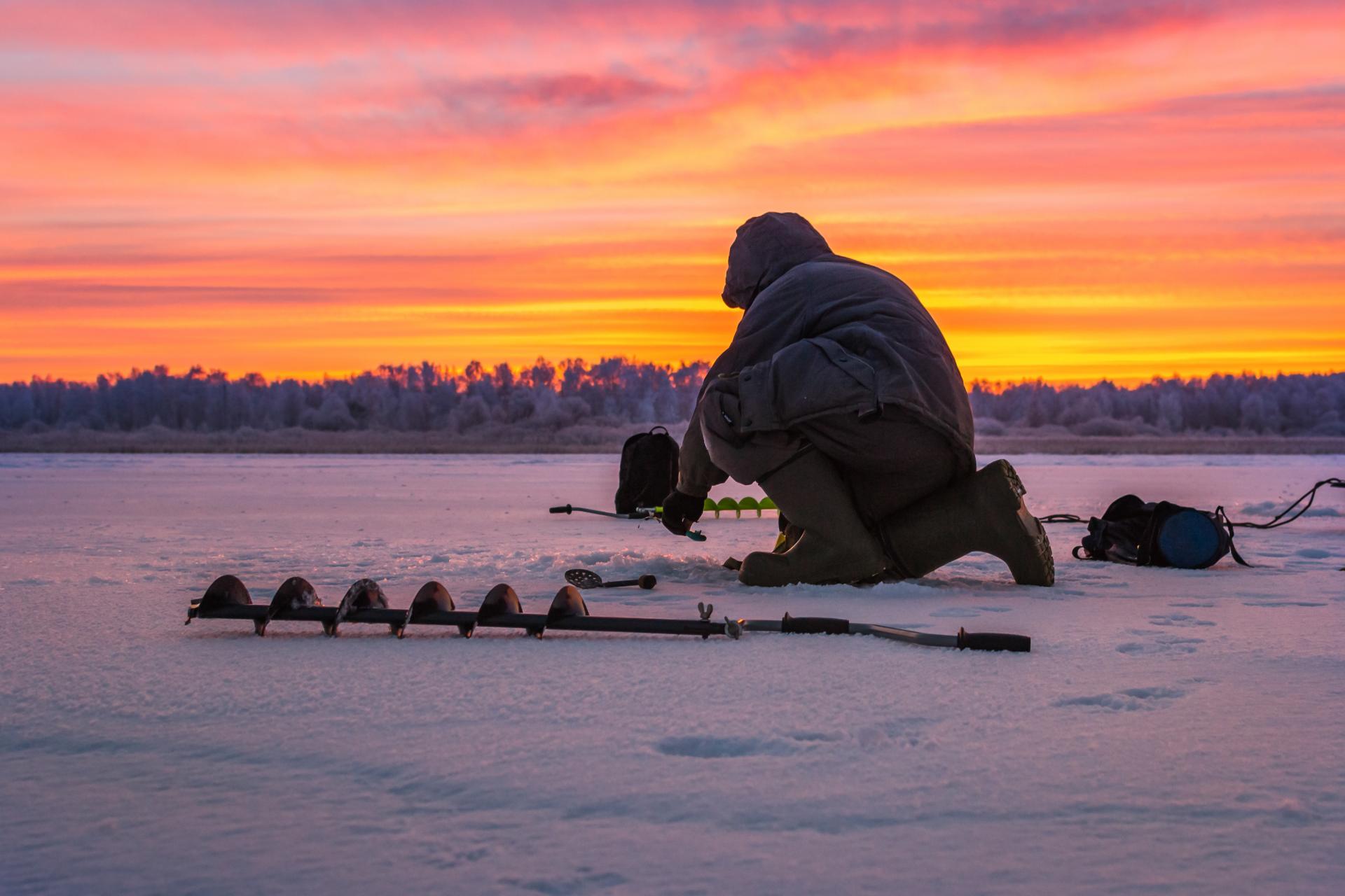 A man fishing outside on the ice in the winter during a sunset